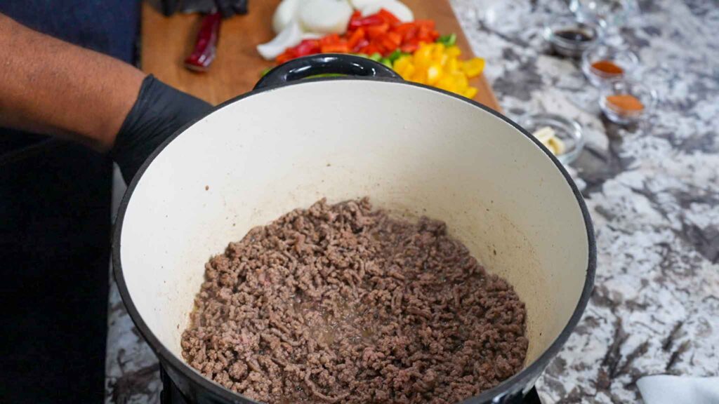 Ground beef browning in a white pot on a marble countertop, perfect for a dirty cabbage recipe. Chopped onions and colorful bell peppers rest on a wooden cutting board, while a person wearing a black glove is visible to the left.