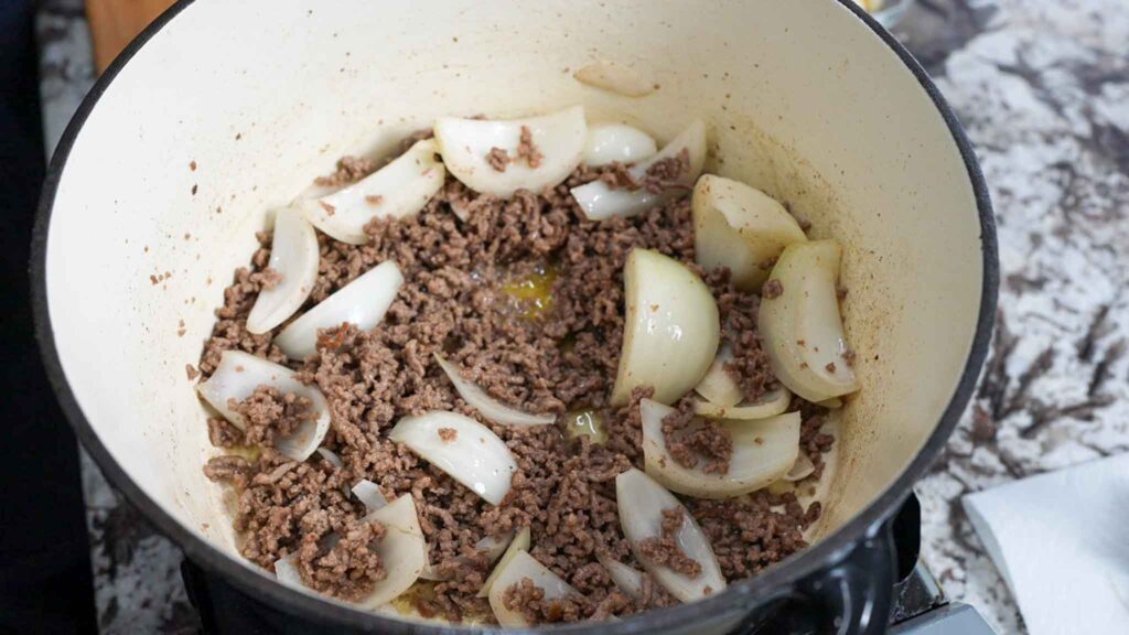 Ground beef and large onion chunks are being cooked together in a white pot on a stove, with some browning visible—just the first step for a hearty dirty cabbage recipe. The pot sits on a marbled countertop.