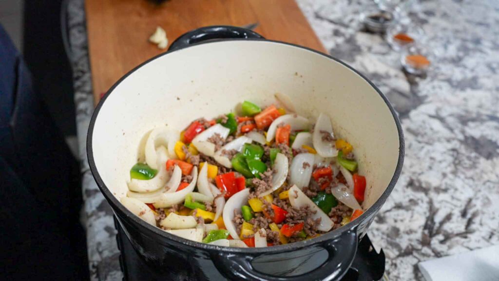 A white Dutch oven sits on a countertop filled with chopped onions, bell peppers, and ground beef—ingredients perfect for starting a dirty cabbage recipe. A wooden cutting board and cooking ingredients are in the background.