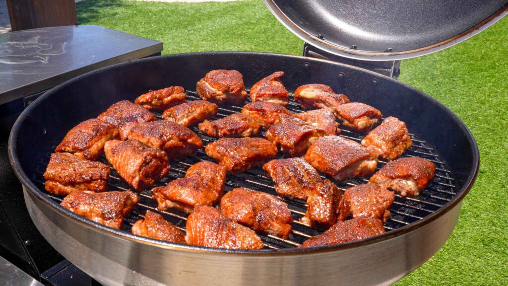 Several pieces of seasoned meat, including Juneteenth BBQ rib tips, are cooking on a round charcoal grill outdoors, with the lid open and green grass visible in the background.
