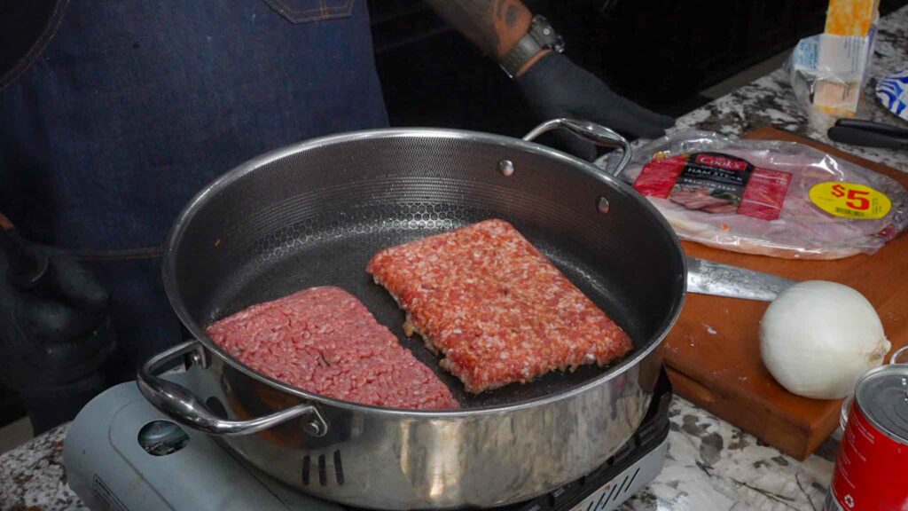 Two rectangular patties of raw ground meat are cooking in a skillet on a stovetop&mdash;ideal for beef sausage burritos. Nearby, there&rsquo;s a cutting board with a whole onion, a can, sliced cheese, and packaged ham marked $5.