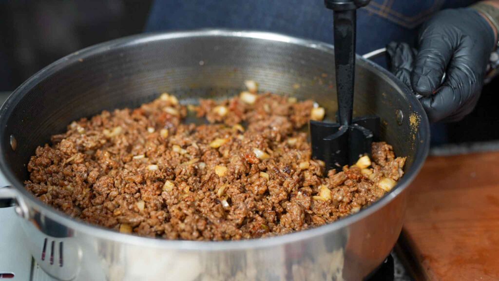 Ground beef and diced onions are being browned and stirred in a large pan with a black spatula by a person wearing a black glove, preparing the savory filling for beef sausage burritos.