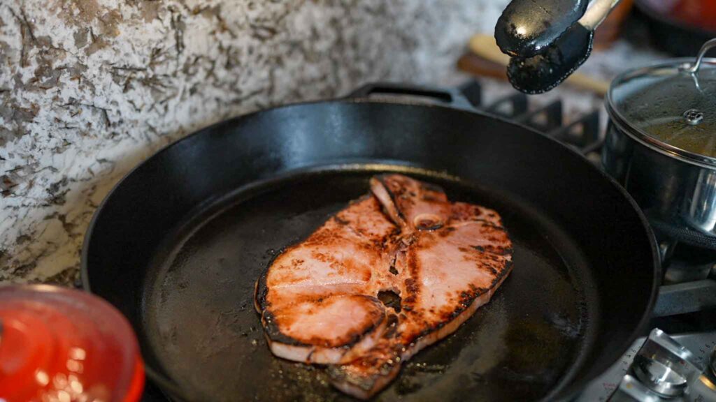 A piece of fish steak is being cooked in a black cast iron skillet on a stovetop, with someone holding metal tongs above it. Nearby, a pot sits on the adjacent burner&mdash;perfect for whipping up sides like beef sausage burritos.