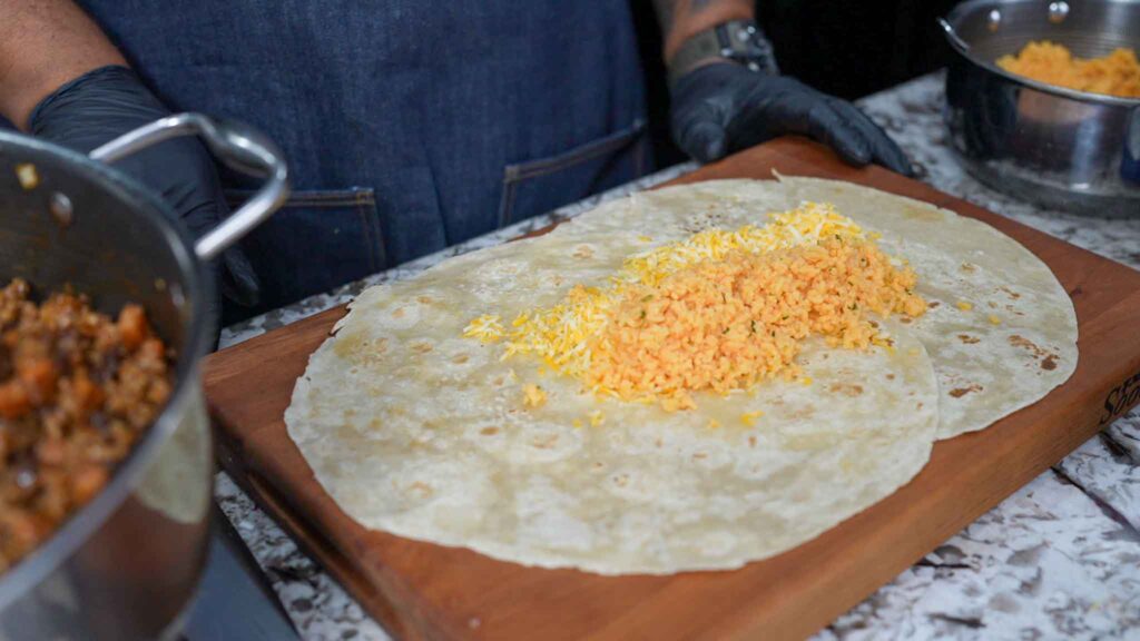 A person wearing black gloves prepares beef sausage burritos by adding shredded cheese and rice to a large tortilla on a wooden board, with pans of filling nearby.