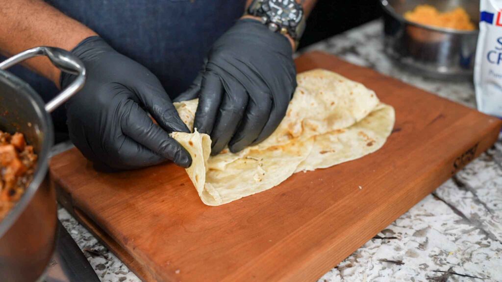 A person wearing black gloves is folding a tortilla on a wooden cutting board, preparing tasty beef sausage burritos, with a pan of food and a bowl of shredded cheese nearby.