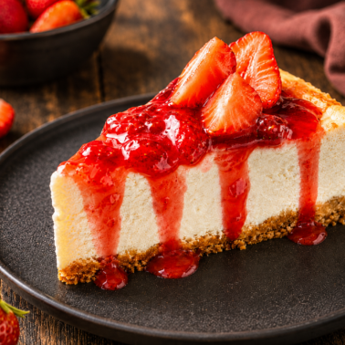 A slice of cheesecake with a graham cracker crust, topped with strawberry sauce and fresh strawberries, served on a black plate. Find this dessert and more in our recipe index, with whole and sliced strawberries in the background.