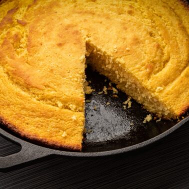 A round skillet of golden cornbread with a wedge slice removed, showing the moist inside. The skillet sits on a dark, textured surface—perfect for adding to your recipe index.