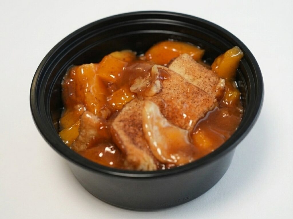 A black plastic bowl filled with peach cobbler from AB&rsquo;s BBQ Fusion food trailer, showing sliced peaches and pieces of golden-brown crust covered in syrupy sauce.
