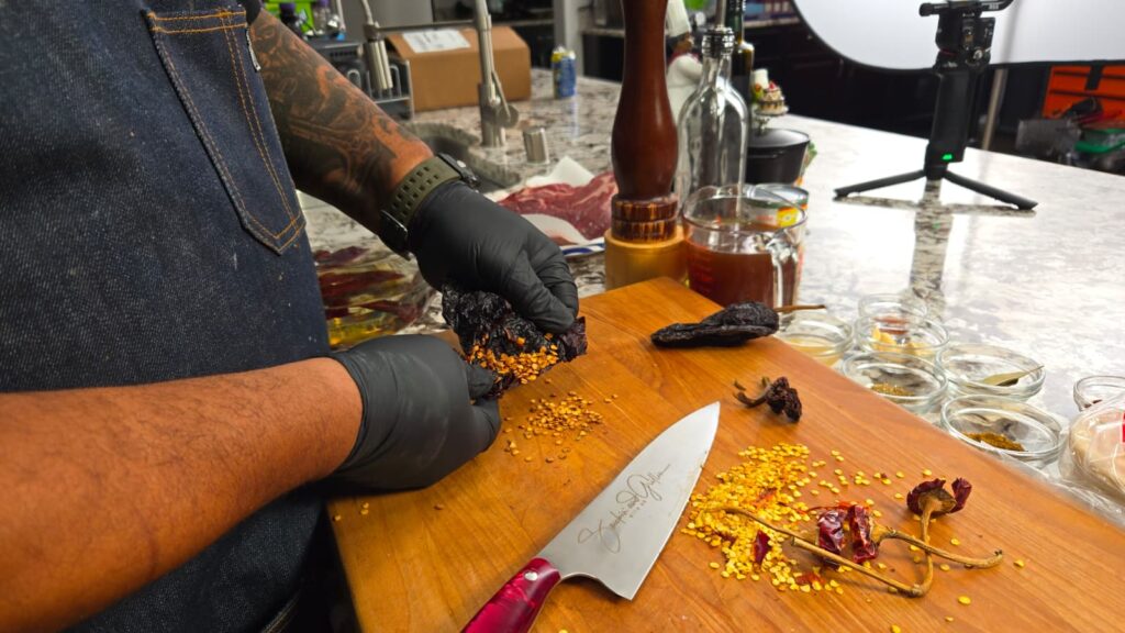 A person wearing black gloves deseeds dried chili peppers on a wooden cutting board. There are scattered seeds, a knife, various spices in small jars, and kitchen items in the background.