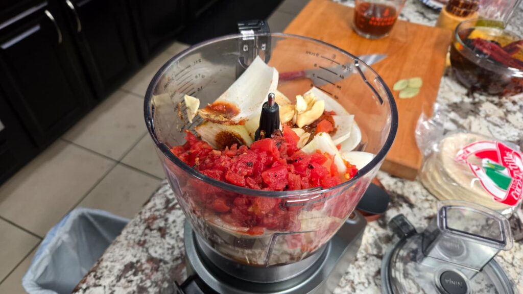 Chopped tomatoes, onions, and garlic cloves in a food processor bowl on a kitchen counter, ready for blending. Nearby are a cutting board, measuring cup, and a package of tortillas.