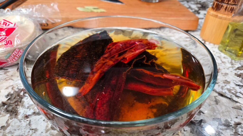 A glass bowl filled with dried red chilies soaking in water sits on a kitchen counter. In the background, a cutting board, tortillas in plastic, and a bottle of oil are partially visible.