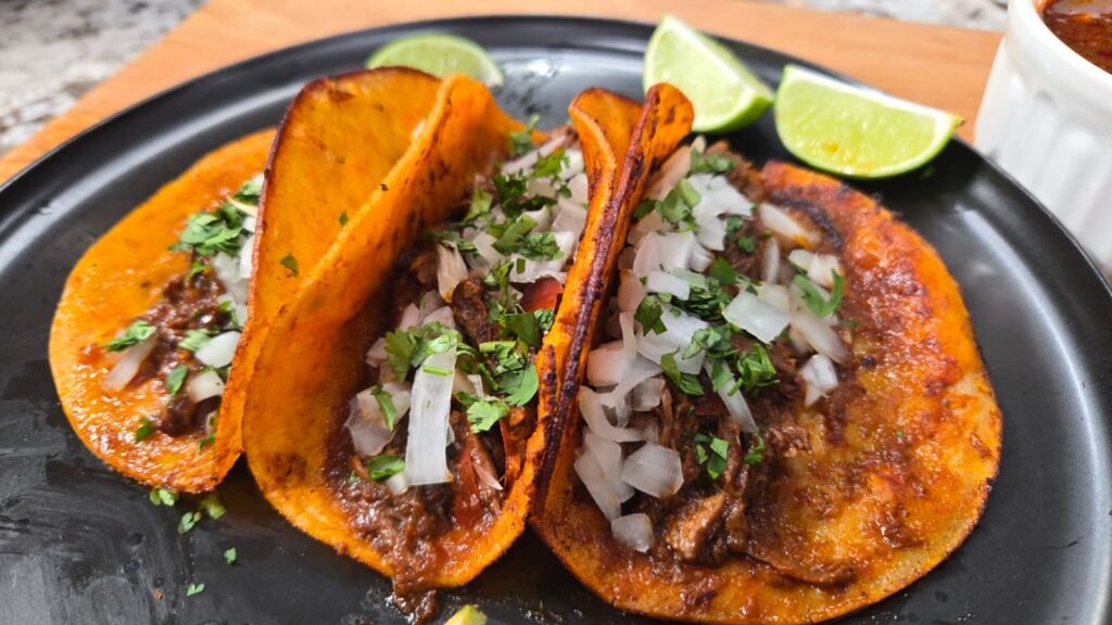 Three birria tacos with orange, crispy tortillas are filled with shredded meat, diced onions, and chopped cilantro, served on a black plate with lime wedges in the background.