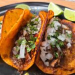 Three birria tacos with orange, crispy tortillas are filled with shredded meat, diced onions, and chopped cilantro, served on a black plate with lime wedges in the background.