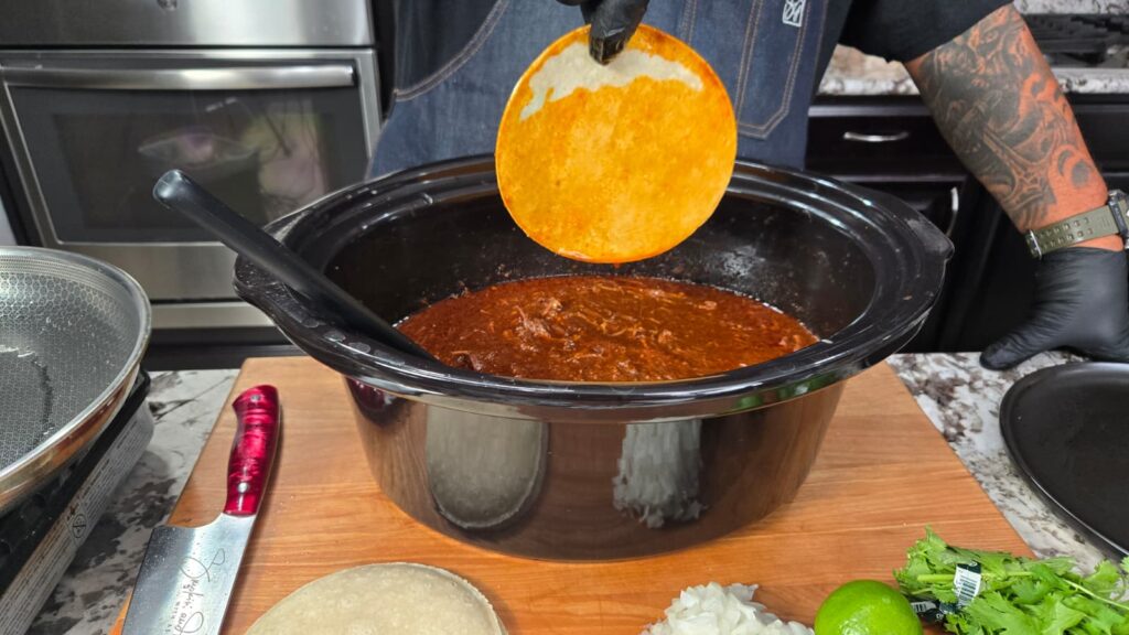 A person dips a tortilla into a slow cooker filled with red birria stew. Chopped onion, cilantro, lime, and a knife are on the wooden counter nearby in a kitchen setting.