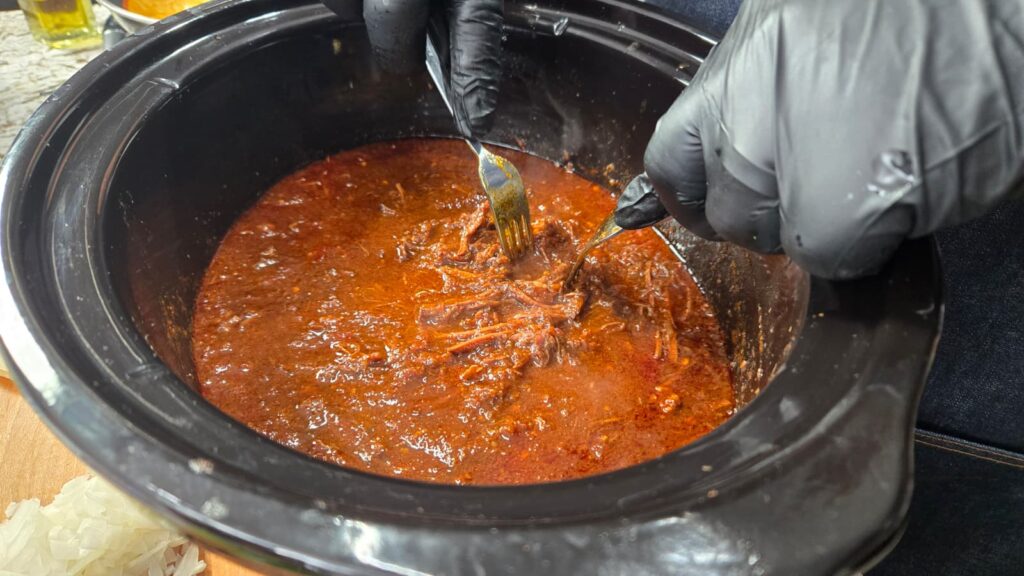 Two gloved hands use forks to shred meat in a slow cooker filled with rich, red sauce. Chopped onions and kitchen items are visible nearby.