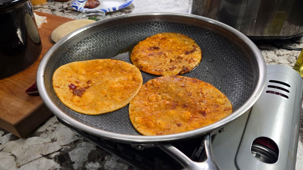 Three corn tortillas frying in a pan with oil on a stovetop, showing a golden, slightly crispy texture. A cutting board and kitchen items are visible in the background.