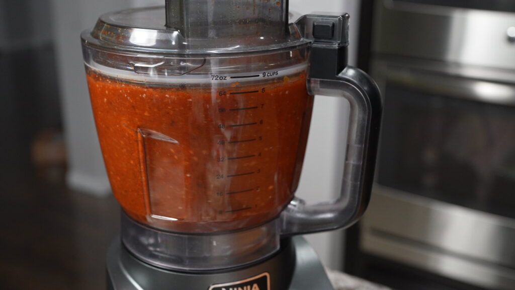 A food processor blending a red sauce mixture, with measurement markings on the container, sits on a kitchen counter next to a stainless steel oven.