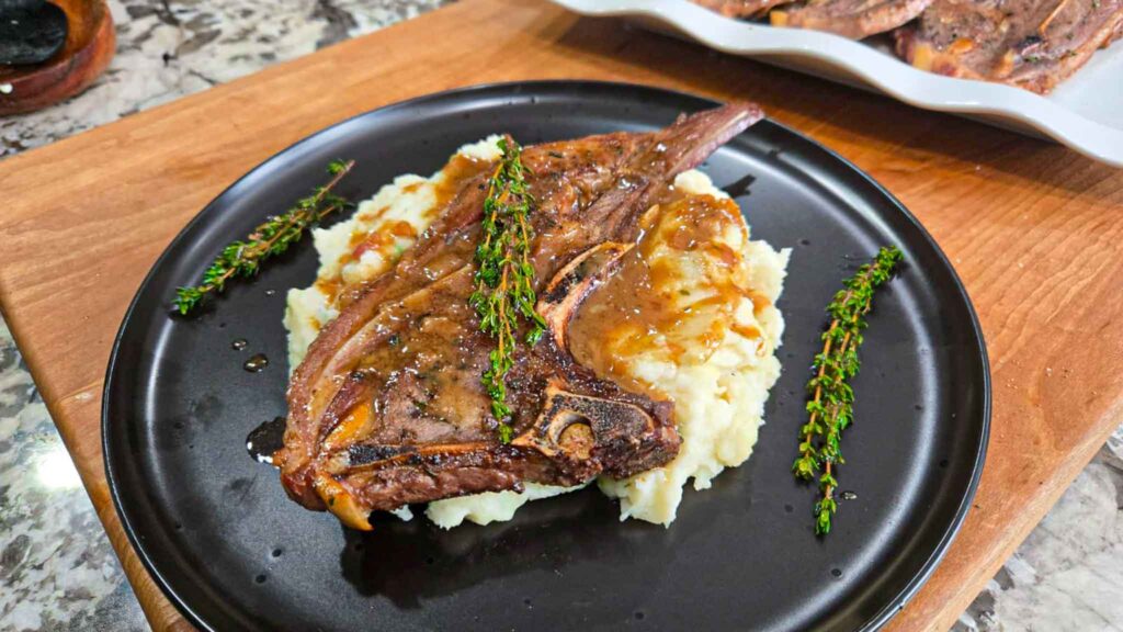 A garlic butter lamb chop with gravy sits on a bed of mashed potatoes, garnished with sprigs of fresh thyme, served on a black plate atop a wooden surface.