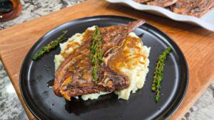 A garlic butter lamb chop with gravy sits on a bed of mashed potatoes, garnished with sprigs of fresh thyme, served on a black plate atop a wooden surface.