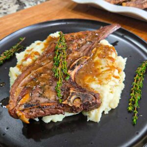 A garlic butter lamb chop with gravy sits on a bed of mashed potatoes, garnished with sprigs of fresh thyme, served on a black plate atop a wooden surface.