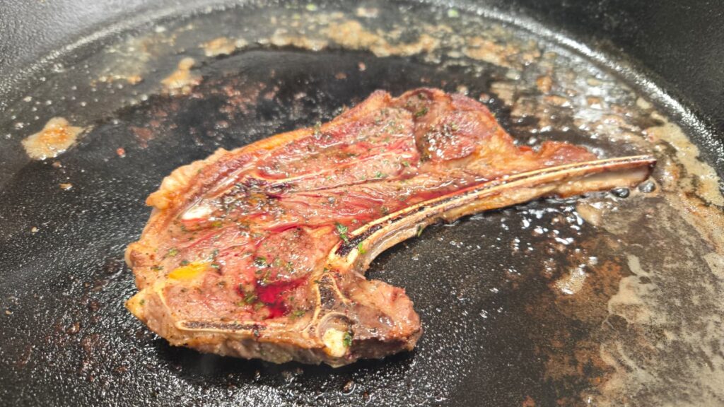 A cooked garlic butter lamb chop with a bone is sizzling in a black frying pan, surrounded by oil and meat juices.