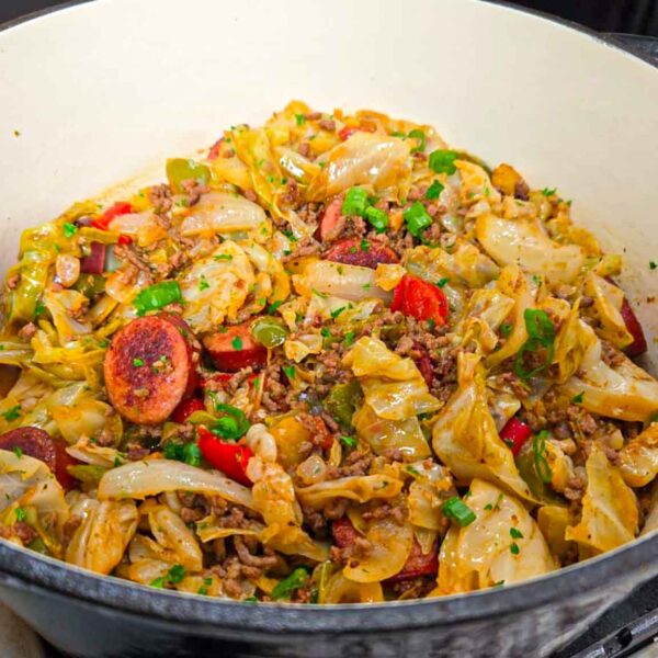 A large pot filled with a hearty dirty cabbage recipe—cooked cabbage, sausage slices, ground meat, bell peppers, and spices—sits on the stovetop with ingredients and utensils nearby.
