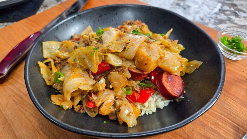 A black bowl filled with cooked cabbage, sliced sausage, ground meat, red bell peppers, and green onions served over rice—a classic dirty cabbage recipe—placed on a wooden board with a knife and a small dish of chopped herbs nearby.