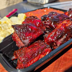 A black tray filled with glazed Juneteenth BBQ rib tips, macaroni and cheese, and baked beans sits on a wooden surface in bright sunlight.