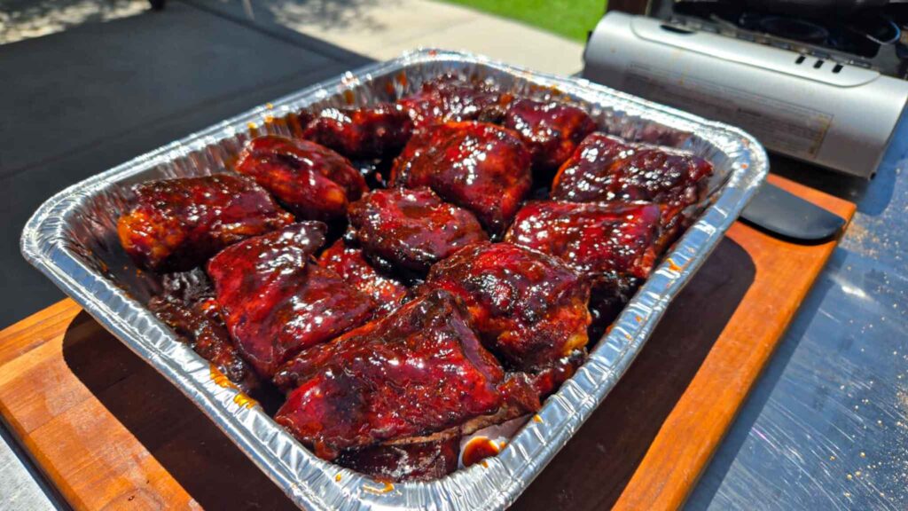 A foil tray filled with glazed Juneteenth BBQ rib tips coated in a rich, dark sauce sits on a wooden board outdoors near a grill.