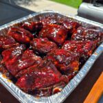 A foil tray filled with glazed Juneteenth BBQ rib tips coated in a rich, dark sauce sits on a wooden board outdoors near a grill.