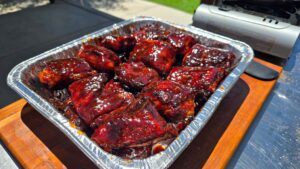 A foil tray filled with glazed Juneteenth BBQ rib tips coated in a rich, dark sauce sits on a wooden board outdoors near a grill.