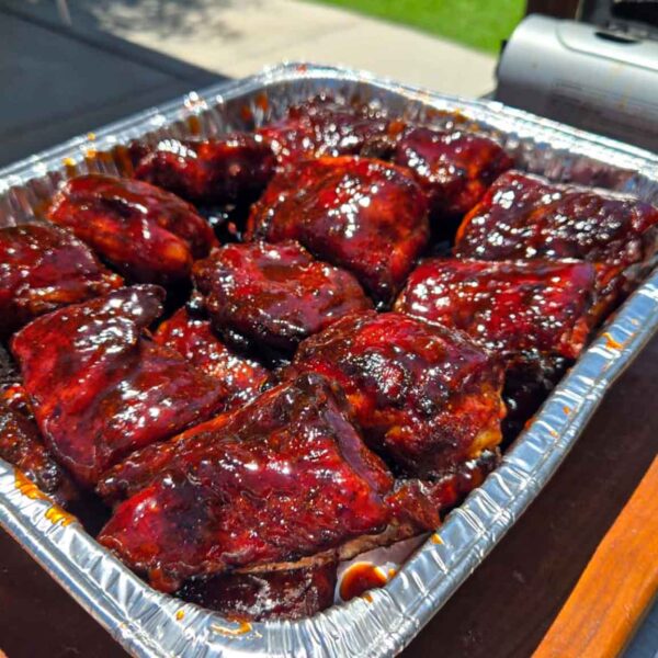 A foil tray filled with glazed Juneteenth BBQ rib tips coated in a rich, dark sauce sits on a wooden board outdoors near a grill.
