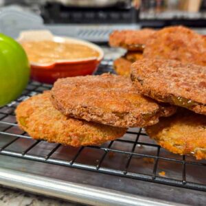 Several golden brown fried green tomato slices rest on a cooling rack. A whole green tomato and a small dish of dipping sauce sit nearby, highlighting the fresh flavor of green tomatoes on a metal tray atop a granite countertop.
