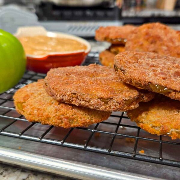 Several golden brown fried green tomato slices rest on a cooling rack. A whole green tomato and a small dish of dipping sauce sit nearby, highlighting the fresh flavor of green tomatoes on a metal tray atop a granite countertop.
