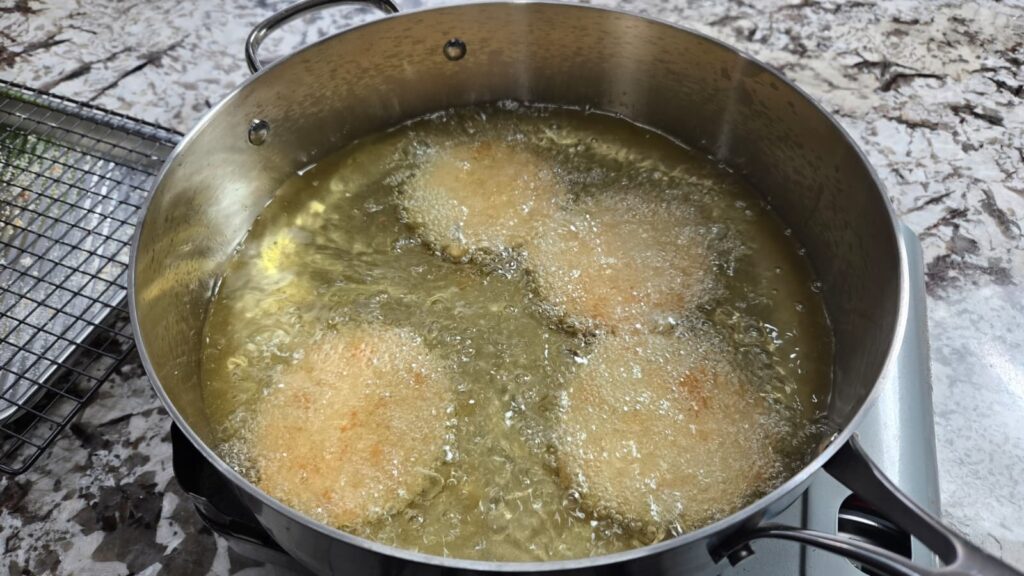 Four breaded green tomatoes are being deep-fried in hot oil in a large metal pot on a stovetop, with bubbling oil surrounding them. A cooling rack sits on the counter nearby.