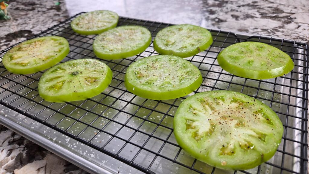 Slices of green tomatoes sprinkled with seasoning are arranged on a wire rack over a baking sheet, set on a marbled countertop.