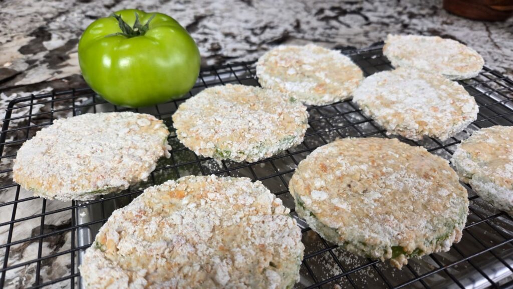 Sliced green tomatoes coated in a breadcrumb mixture rest on a cooling rack, ready for frying. A whole green tomato sits next to the slices, highlighting the freshness of these vibrant green tomatoes on a marble countertop.