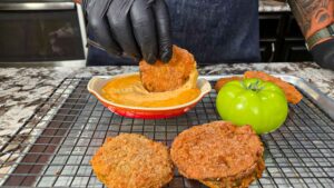 A person wearing black gloves dips a fried green tomato slice into a creamy sauce. Crispy fried green tomatoes rest on a cooling rack beside a fresh green tomato, all on a kitchen counter.