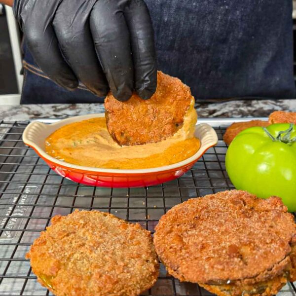 A person wearing black gloves dips a fried green tomato slice into a creamy sauce. Crispy fried green tomatoes rest on a cooling rack beside a fresh green tomato, all on a kitchen counter.