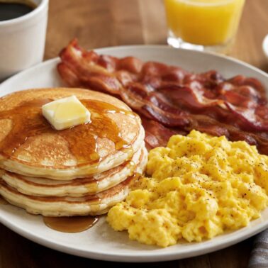 A plate of breakfast food with pancakes topped with butter and syrup, scrambled eggs, and crispy bacon. In the background are a cup of coffee, a glass of orange juice, and a small dish of butter from our recipe index.