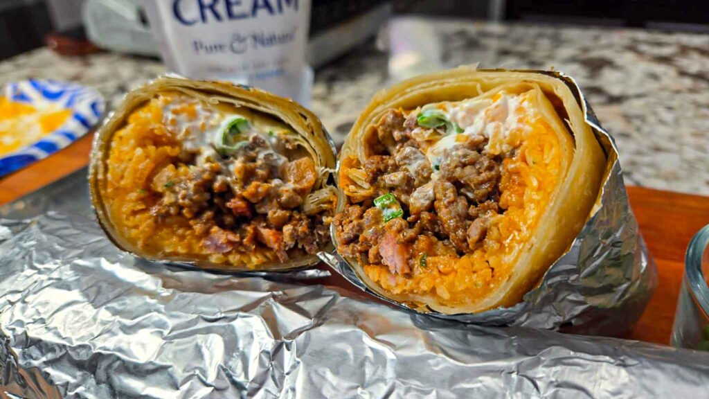 A close-up of beef sausage burritos cut in half, revealing seasoned meat, rice, and sour cream inside a tortilla. The burritos are wrapped in foil and placed on a counter with a cup of cream in the background.