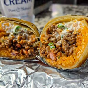 A close-up of beef sausage burritos cut in half, revealing seasoned meat, rice, and sour cream inside a tortilla. The burritos are wrapped in foil and placed on a counter with a cup of cream in the background.