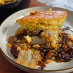 A close-up of a plate with a serving of casserole topped with golden cornbread, ground meat, and dirty cabbage, with a fork resting on the side. A cast iron skillet with more casserole is in the background.