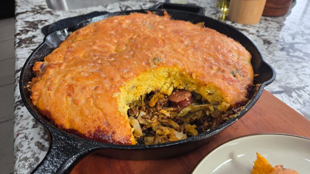 A dirty cabbage cornbread skillet sits on a wooden board, its golden crust partially sliced to reveal a savory filling of sausage, vegetables, and tender cabbage on a marble countertop.