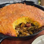 A dirty cabbage cornbread skillet sits on a wooden board, its golden crust partially sliced to reveal a savory filling of sausage, vegetables, and tender cabbage on a marble countertop.