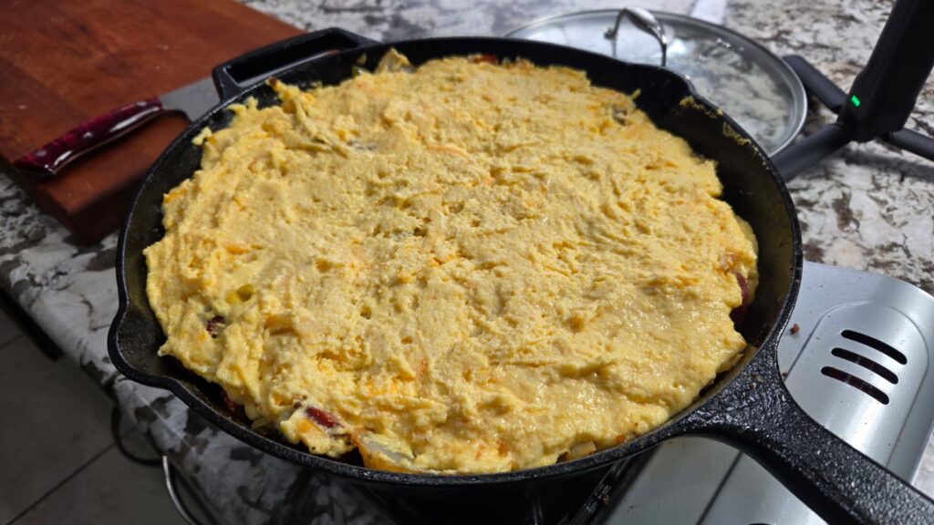 A dirty cabbage cornbread skillet sits on the stovetop, filled with thick, yellow batter ready to be baked. The speckled countertop holds a cutting board and kitchen utensils in the background.