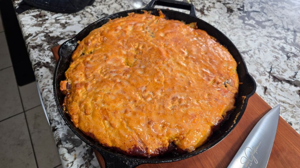 A golden-brown dirty cabbage cornbread skillet sits on a speckled countertop next to a silver knife on a wooden board. The crispy topping covers a savory fruit filling underneath.