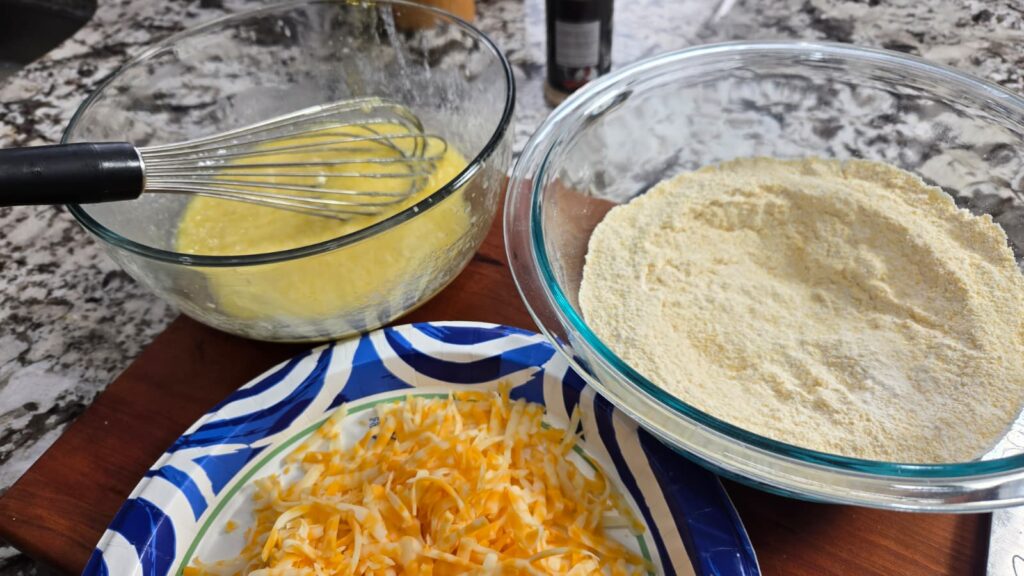 A mixing bowl with whisked wet ingredients, a bowl of dry cornmeal mixture, and a plate of shredded yellow and white cheese sit on the countertop&mdash;classic prep for a dirty cabbage cornbread skillet, ready for baking.