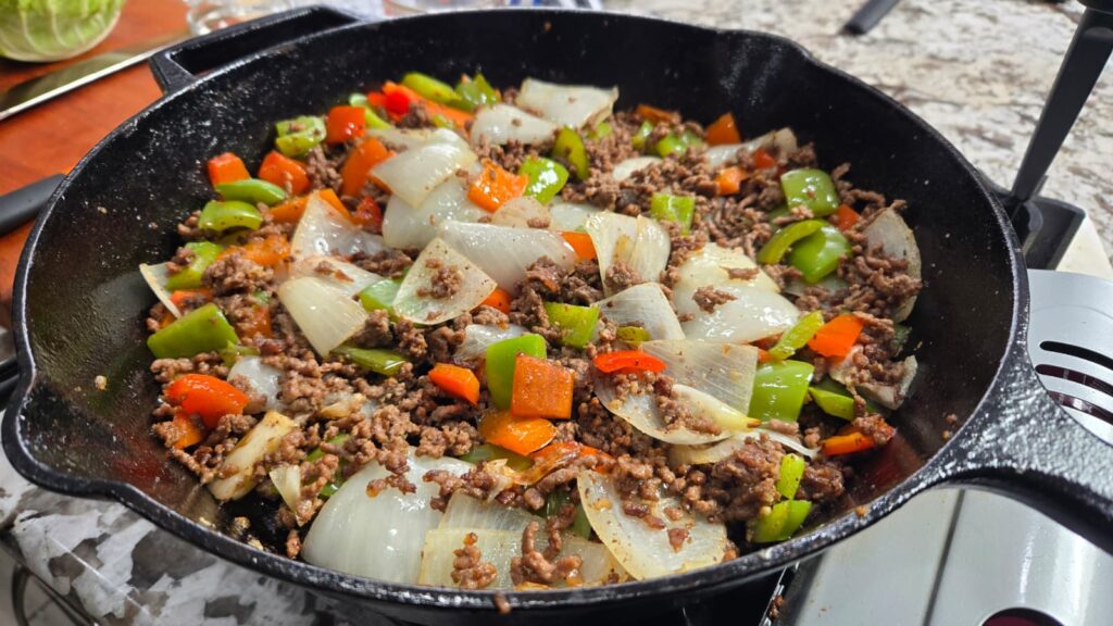 A cast iron skillet on a stovetop filled with saut&eacute;ed ground beef, chopped onions, and green and red bell peppers&mdash;a colorful, hearty dish reminiscent of a classic dirty cabbage cornbread skillet.