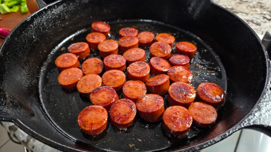 Sliced sausage pieces are being pan-fried in a black cast iron skillet, with some browning visible. The sausages, arranged in a single layer and cooking in oil, evoke the comforting aroma of a classic dirty cabbage cornbread skillet meal.
